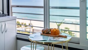 Une petite table ronde sur laquelle sont posés un magazine, une miche de pain et une plante, près d’une fenêtre donnant sur une plage.