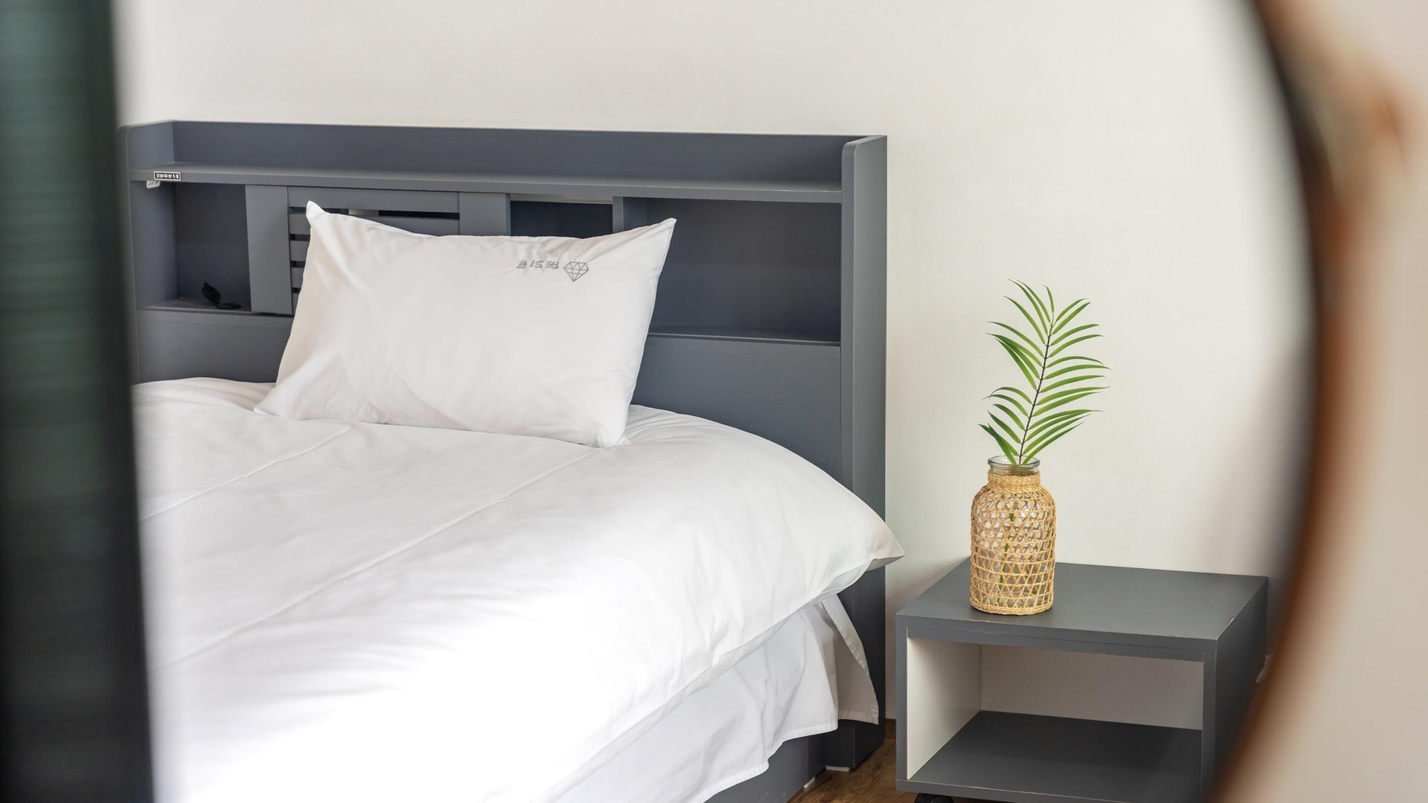 Ein Bett mit weißer Bettwäsche und einem grauen Kopfteil, ein kleiner Nachttisch mit einer gewebten Vase, in der eine Pflanze steht, und ein grauer Beistelltisch.