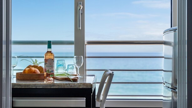Une table avec une bouteille de Mouton Cadus 2022, un panier de pain et des verres à vin, le tout placé sur un balcon donnant sur la mer.
