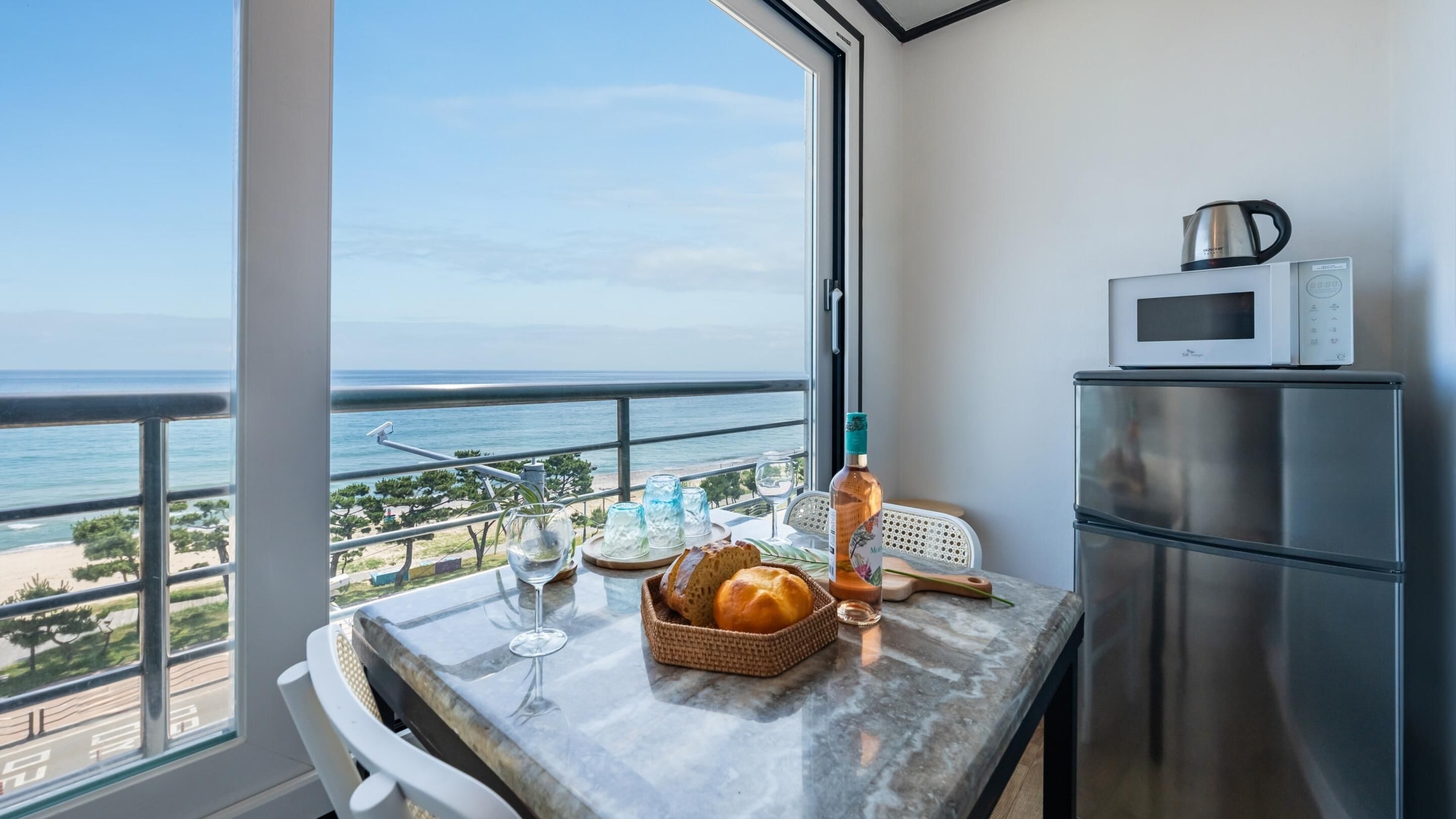 A balcony with a view of the sea, a table with a bottle, glasses, and bread, and a kitchen area with a microwave and refrigerator.