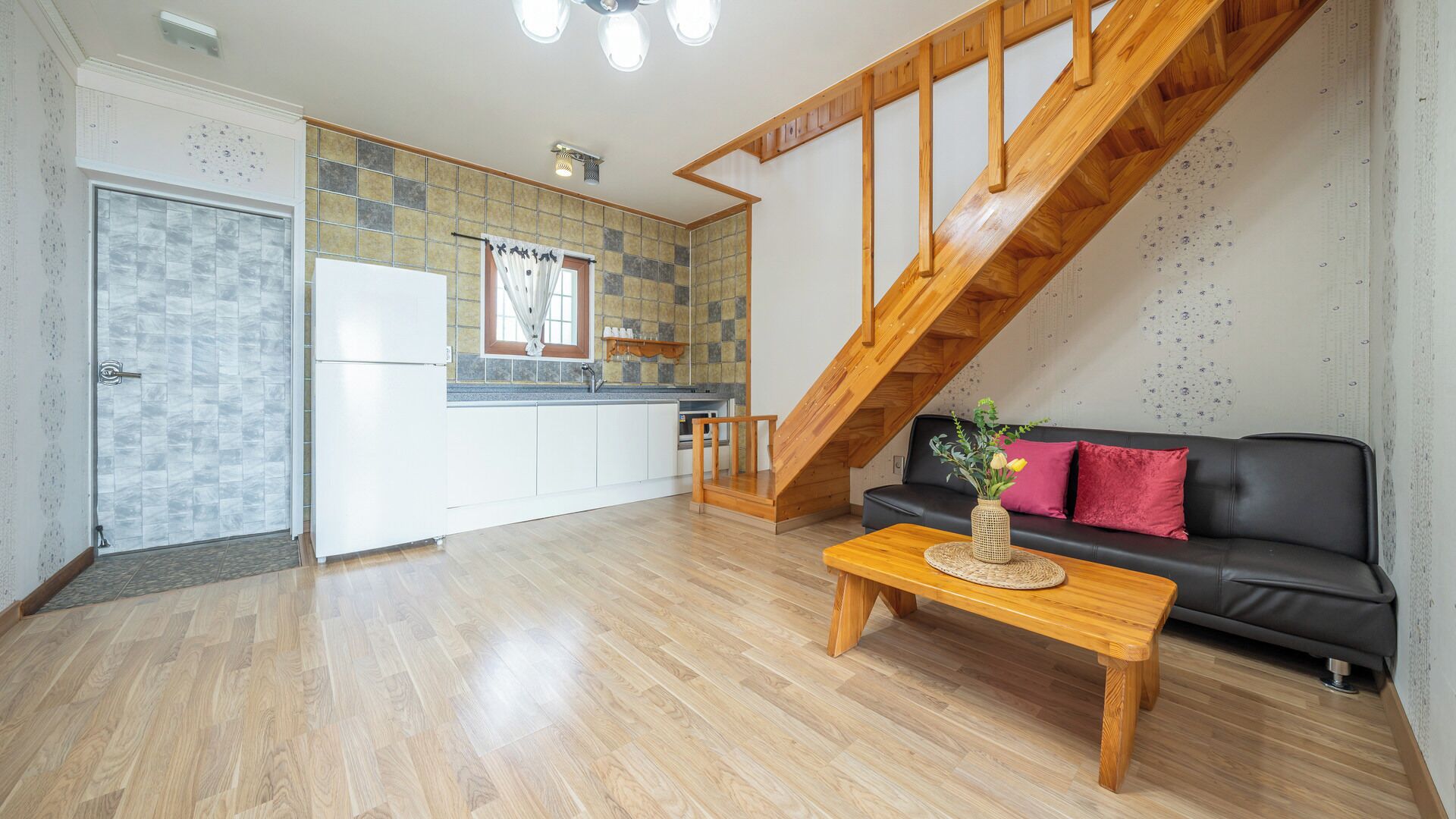 A modern kitchen with white cabinets, a wooden staircase, a black sofa with red cushions, and a wooden coffee table.