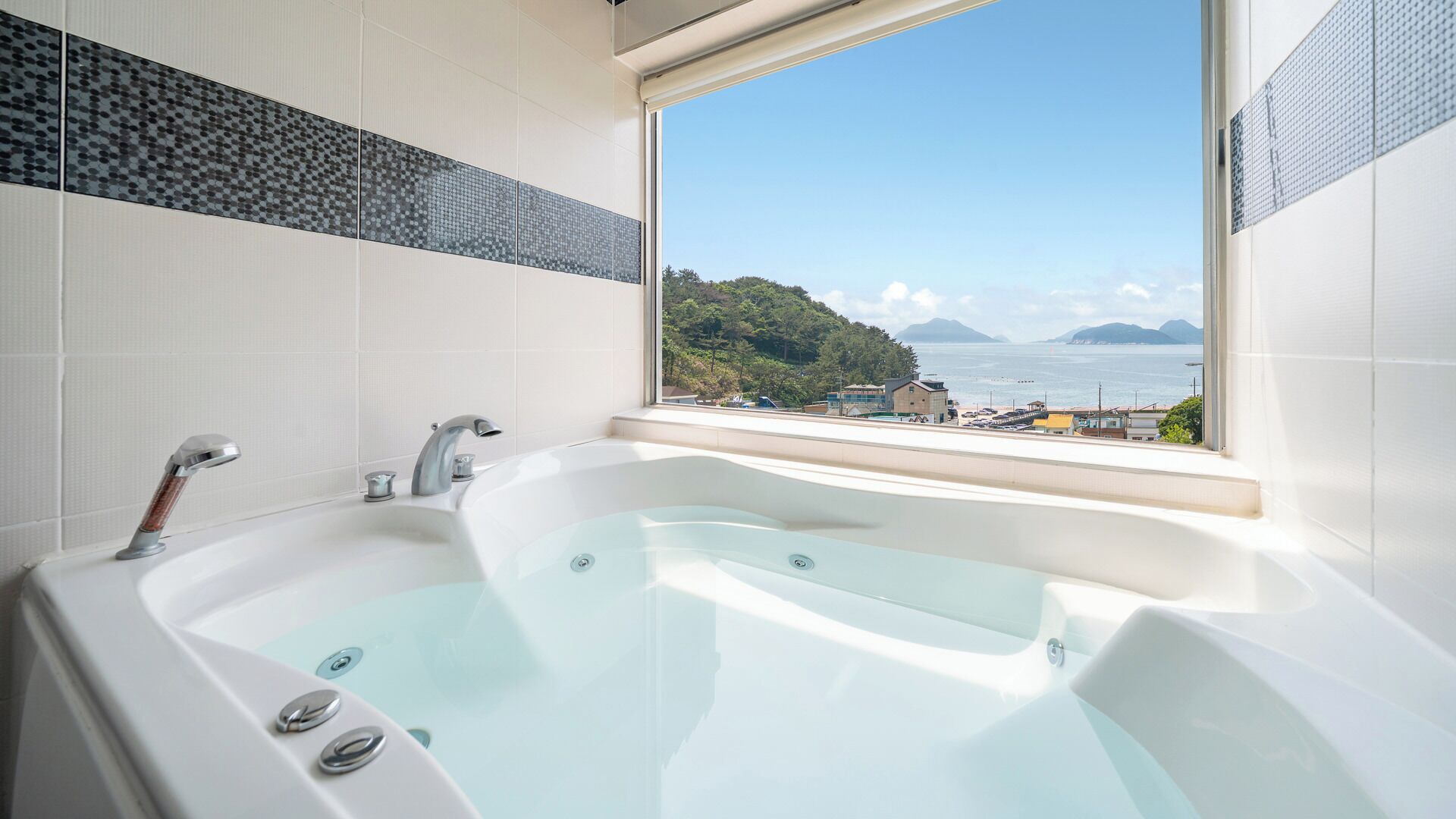 A bathroom with a large bathtub, a window with a view of a coastal landscape, and tiled walls.