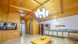A spacious living room with a wooden ceiling, a television mounted on the wall, a wooden coffee table with fruit, and a kitchen area with cabinets and a sink. - Geoje Gaonara Pension (Geoje)
