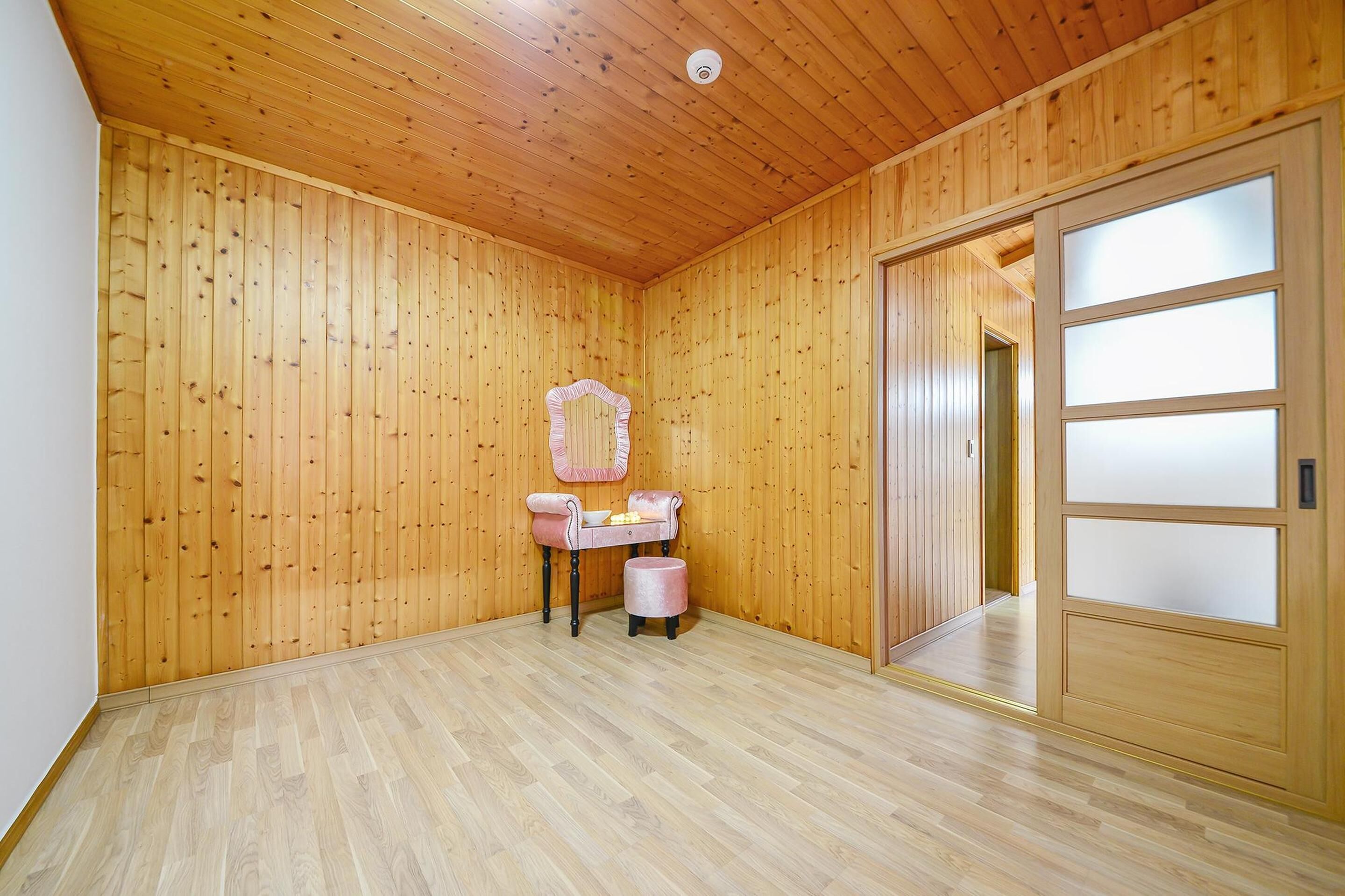 A room with wooden walls and ceiling, a wooden floor, a vanity with a mirror, and a sliding door with frosted glass panels.