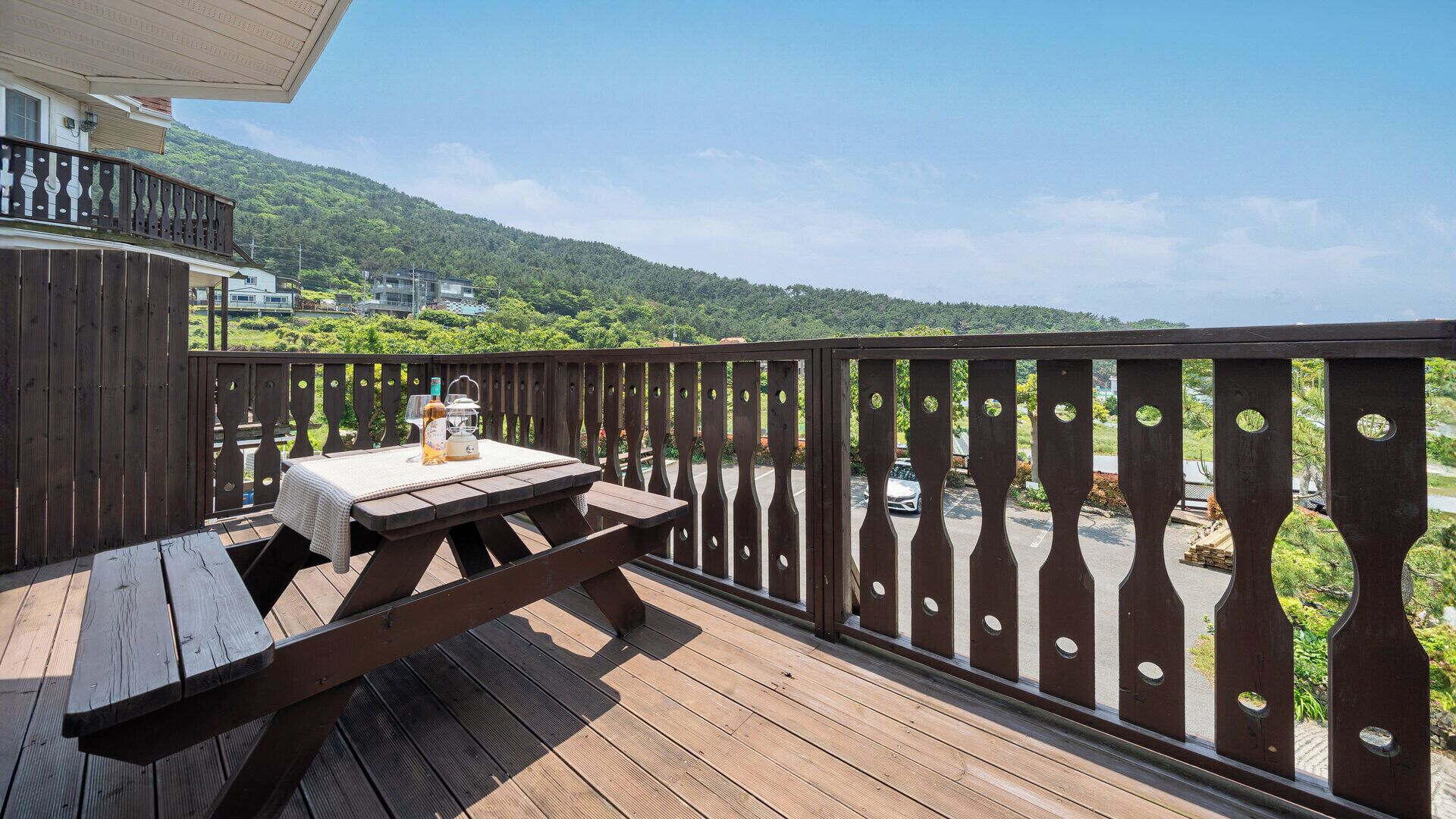 Um deck de madeira com uma mesa de piquenique e um candeeiro, com vista para uma paisagem verdejante e uma estrada.