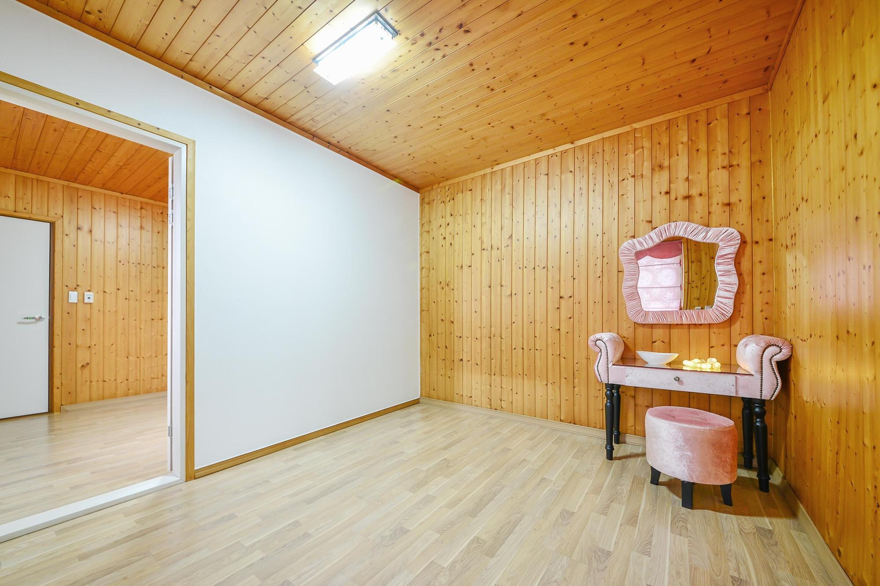 A room with wooden walls and ceiling, a white door, a vanity with a mirror, and a small stool.