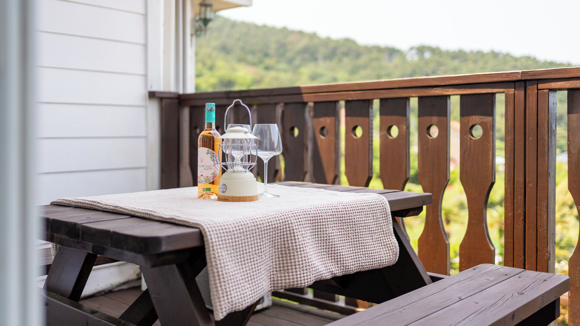 Une terrasse en bois avec une table dressée pour deux personnes, agrémentée d’une bouteille et de verres, ainsi que d’une lanterne.