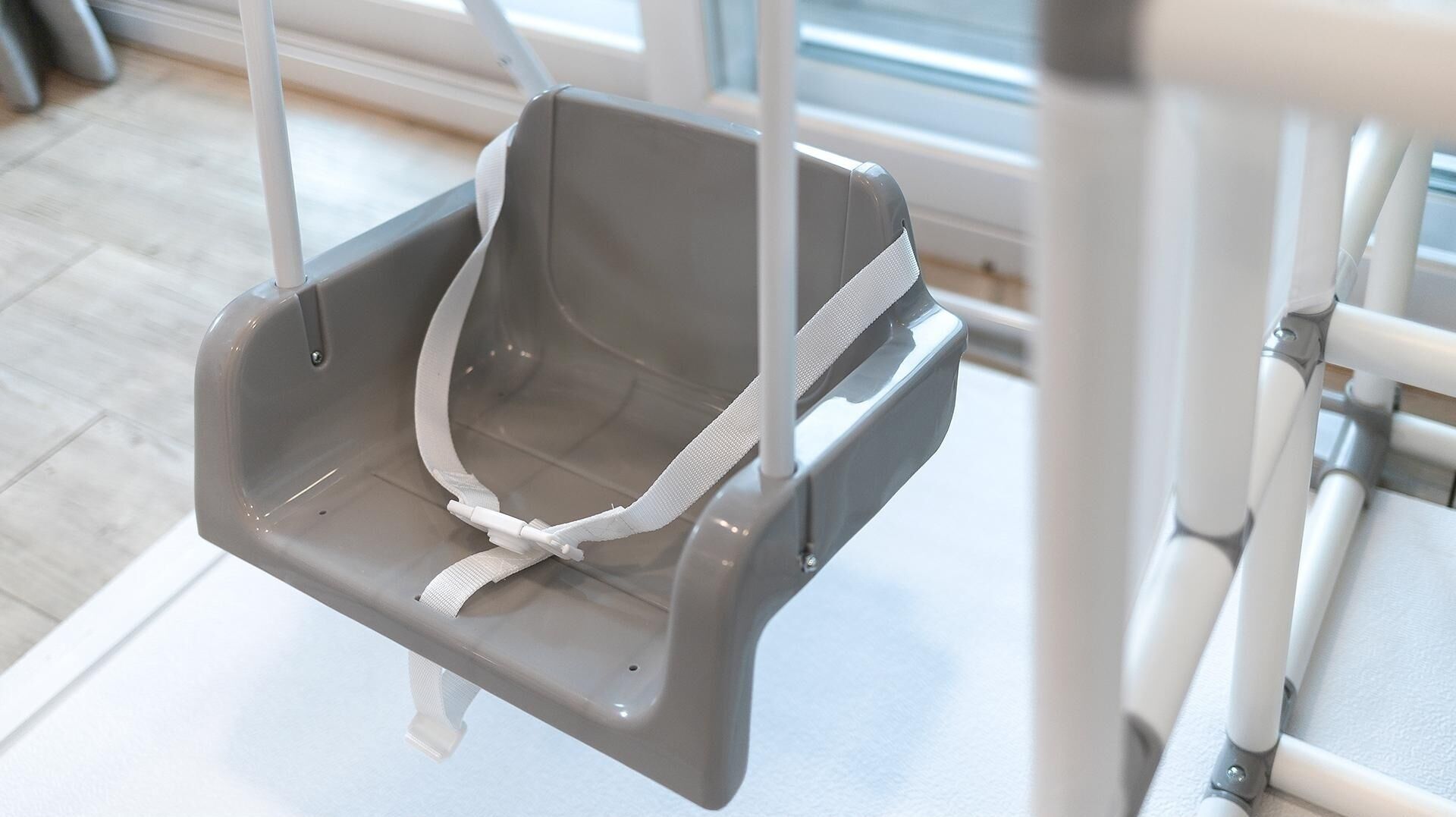 A gray baby swing suspended from a white metal frame, with a white safety harness attached.