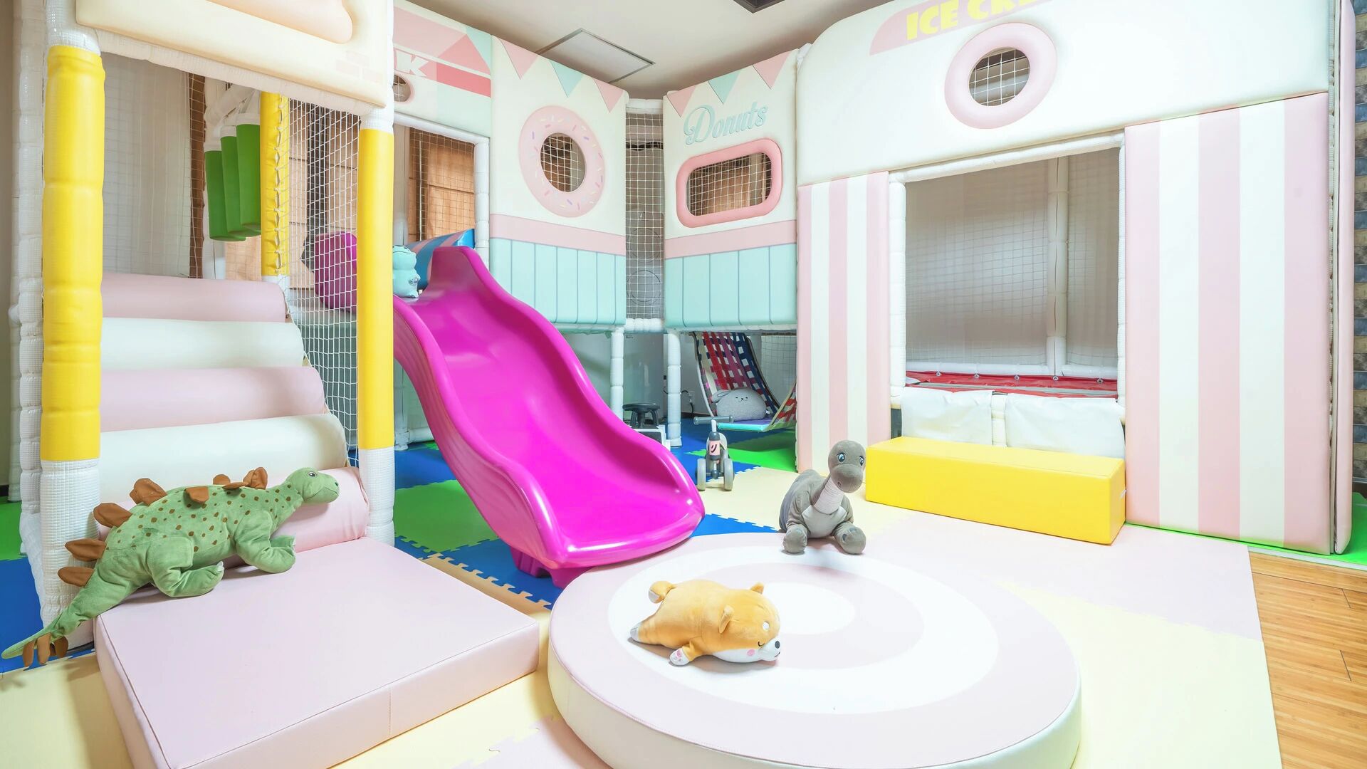 A children's play area with a slide, soft play structures, and a circular seating area with a plush toy.