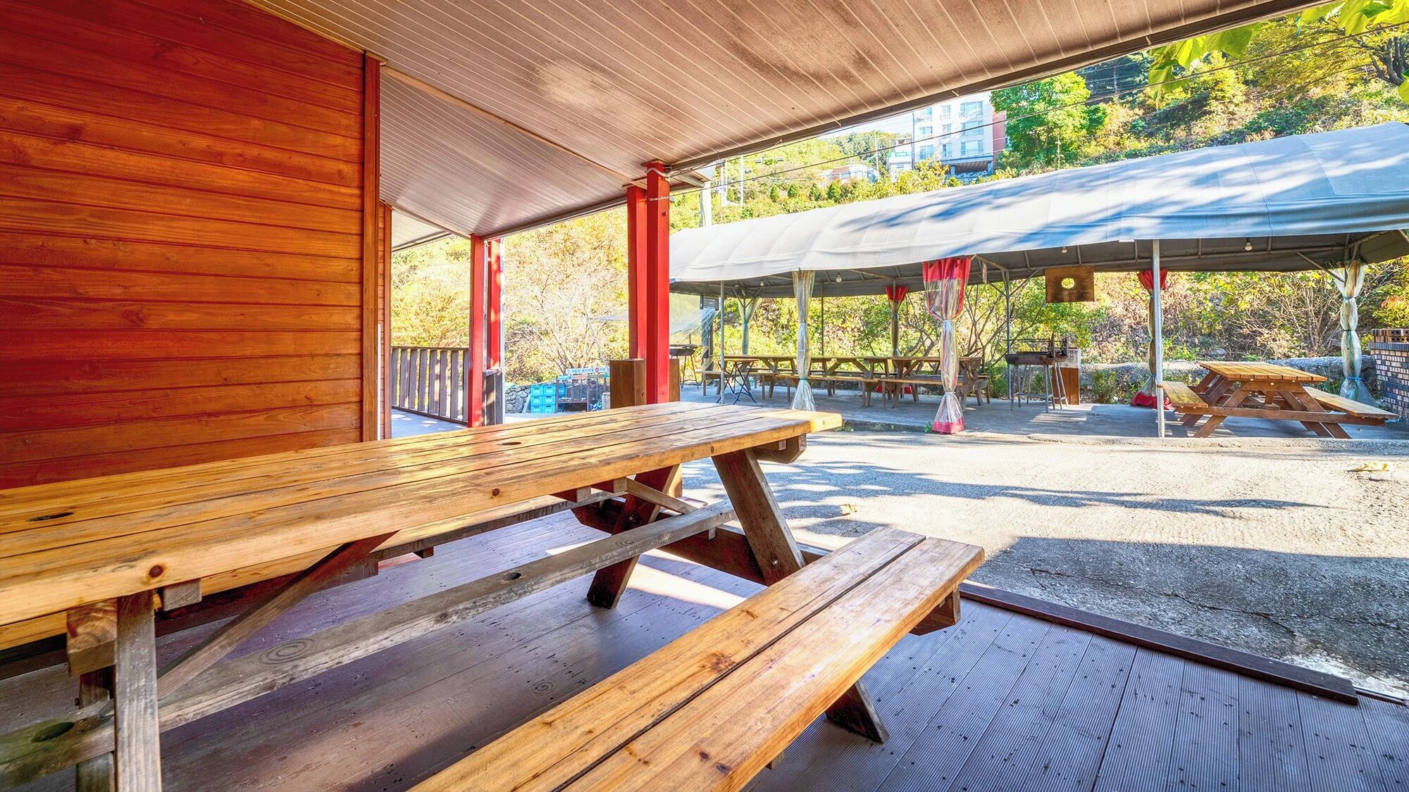 A covered picnic area with wooden benches and tables, a red column, and a view of trees and buildings in the background.