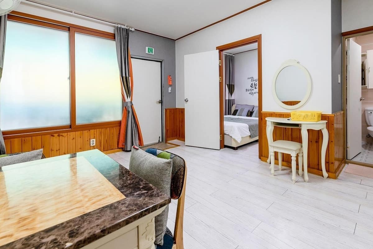 A hotel room with a dining table, a small white table with a mirror and basket, a bed, and a bathroom with a toilet and sink.