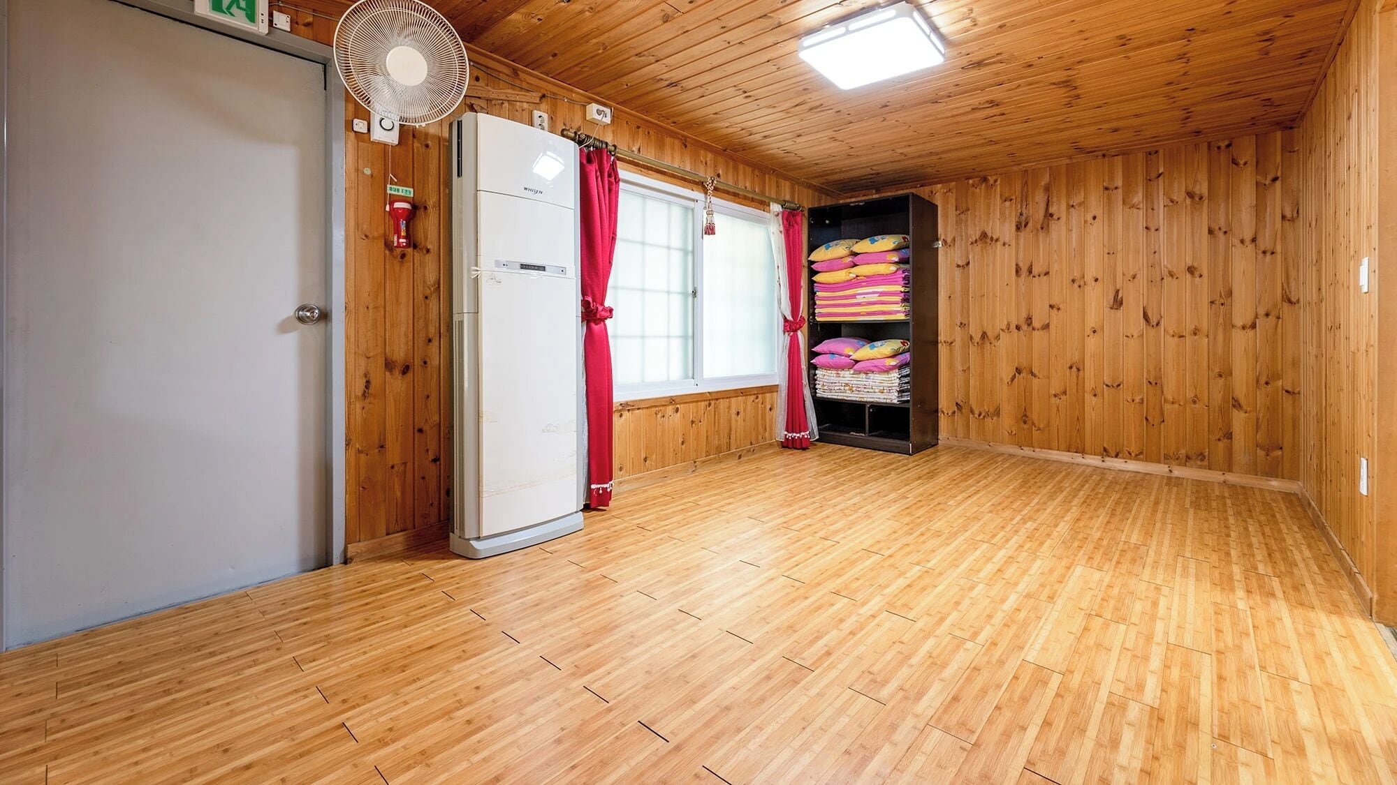 Une pièce aux murs et au plancher en bois, avec une porte blanche, une fenêtre ornée de rideaux rouges, un ventilateur et une étagère sur laquelle sont pliés des vêtements.