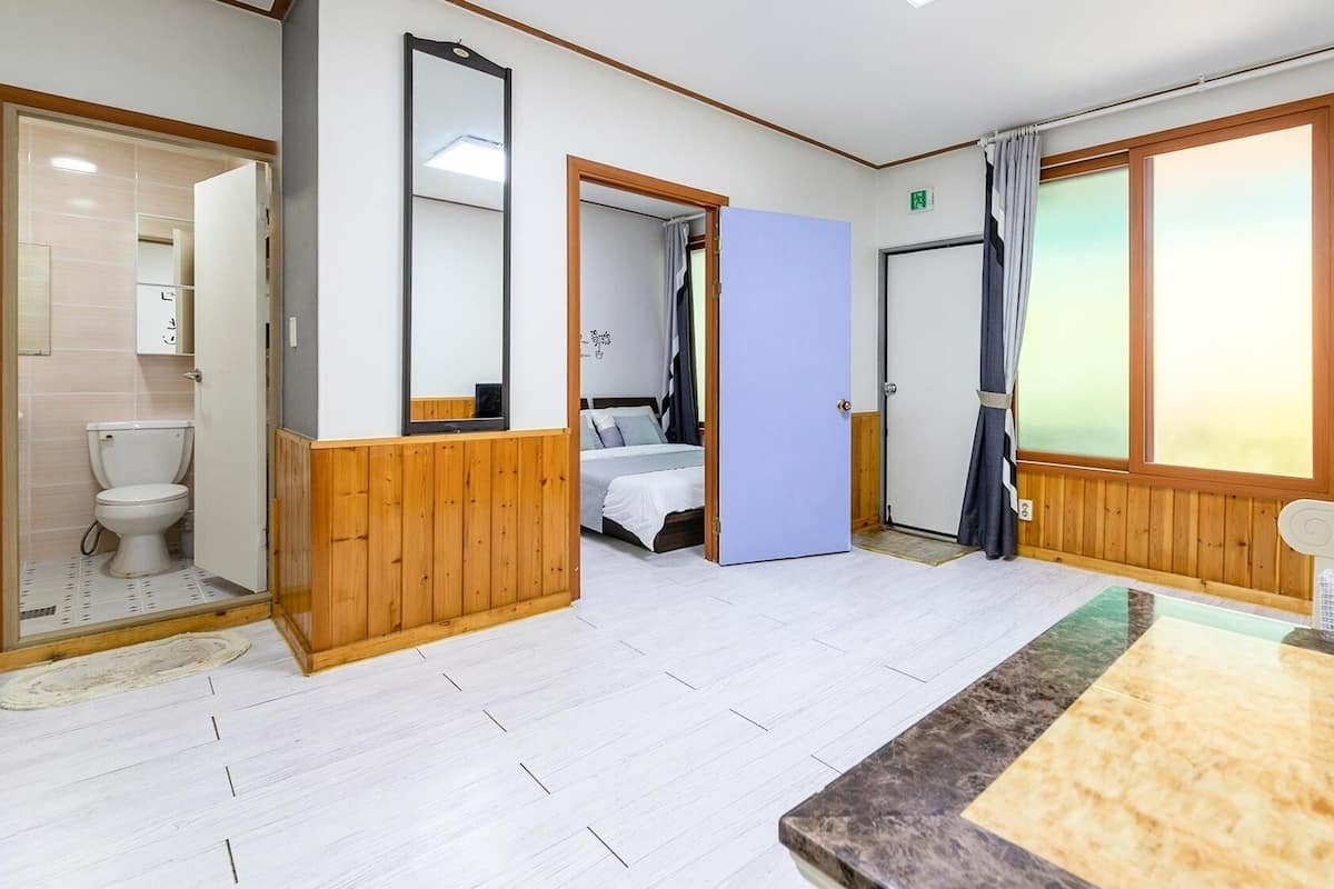 A hotel room with a wooden floor, a bed, a bathroom with a toilet, and a large window with curtains.