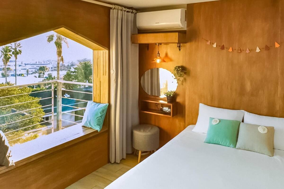 A hotel room with a large bed, a window with a view of palm trees, a small round table, and a mounted air conditioner.