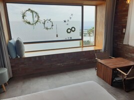 Quarto com uma grande janela adornada com guirlandas decorativas e desenhos de estrelas. Uma escrivaninha e uma cadeira de madeira estão posicionadas perto da janela, e um banco estofado se encontra em primeiro plano.