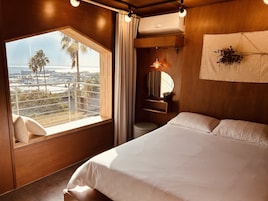 Un dormitorio con una cama grande, una ventana con vistas a palmeras y edificios, una mesita de noche con lámpara, y un aire acondicionado instalado en la pared.