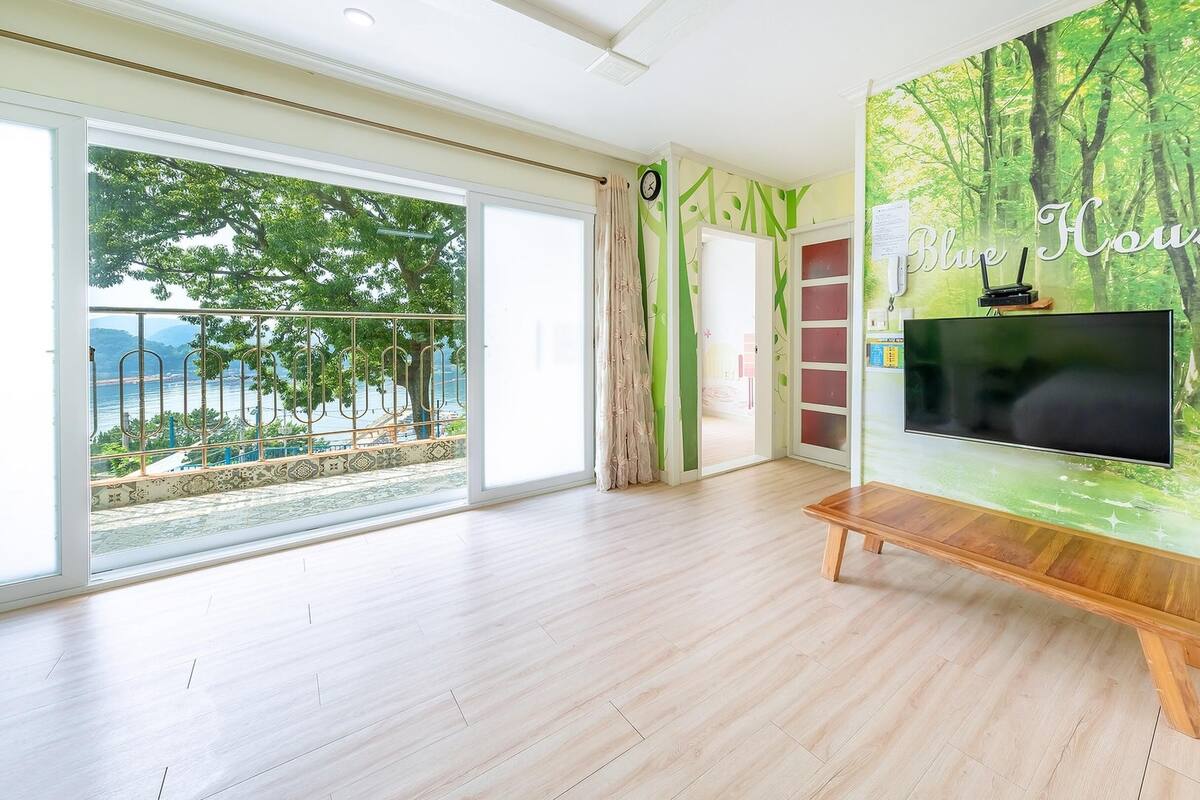A spacious room with a large window, a flat-screen TV mounted on a wooden stand, and a wall mural depicting a forest scene.