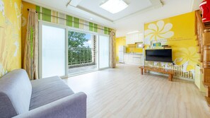 A living room with a sofa, a TV on a wooden stand, and a balcony with a view of trees. - Tongyoung Blue House Spa Pension (Tongyeong)