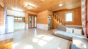A spacious living area with a wooden ceiling, a kitchen with white cabinets, a sofa with patterned cushions, and a staircase leading to an upper level. - Tongyoung Blue House Spa Pension (Tongyeong)