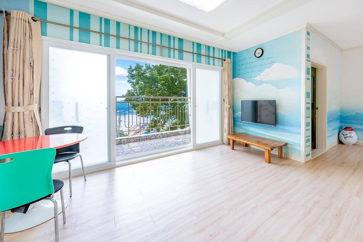 A spacious room with a large sliding glass door leading to a balcony, a wooden bench, a TV mounted on the wall, and a small table with chairs.