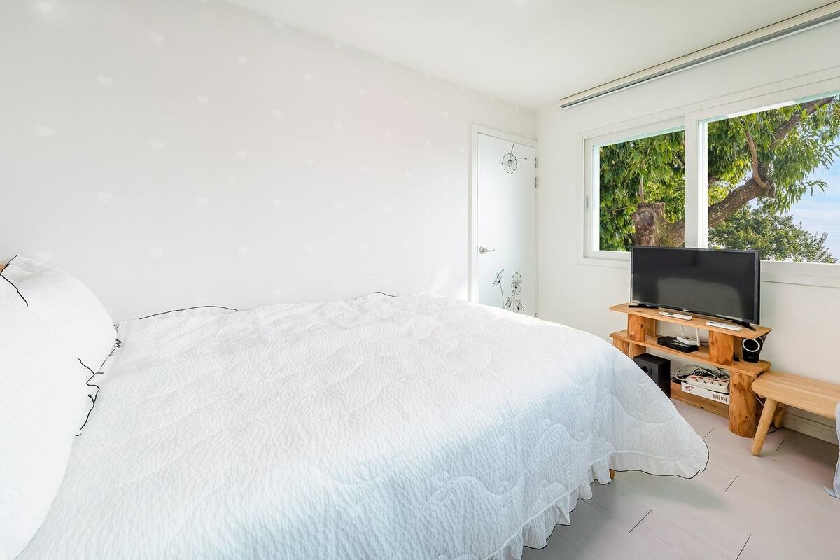 A bedroom with a large bed, a TV on a wooden stand, and a window with a tree view.