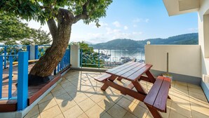 A terrace with a wooden table and benches, overlooking a marina and hills. - Tongyoung Blue House Spa Pension (Tongyeong)