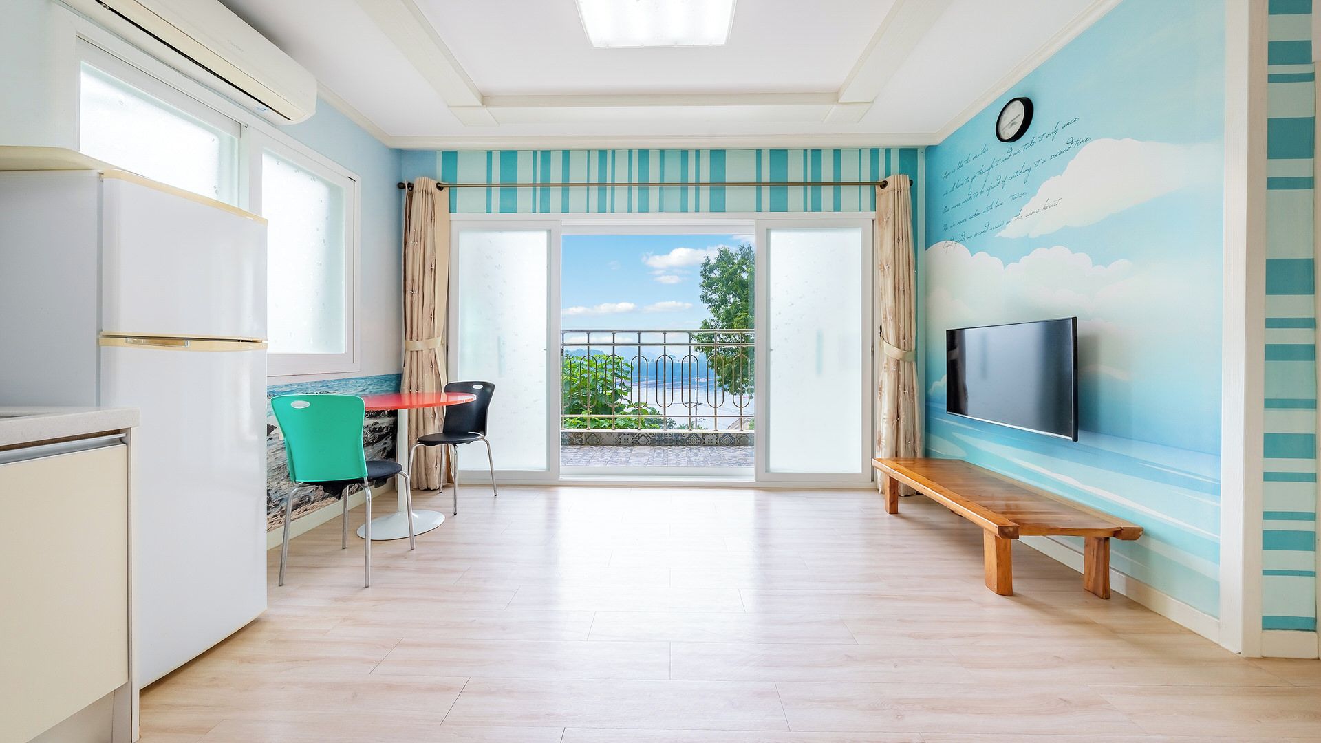 Una habitación con cocina, comedor con sillas, un banco, televisor montado en la pared y balcón con vistas.