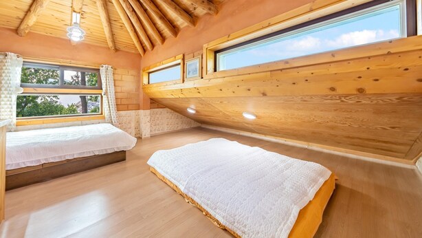 A cozy room with two beds, wooden ceiling beams, and large windows offering a view of the outdoors. - Tongyoung Blue House Spa Pension (Tongyeong)