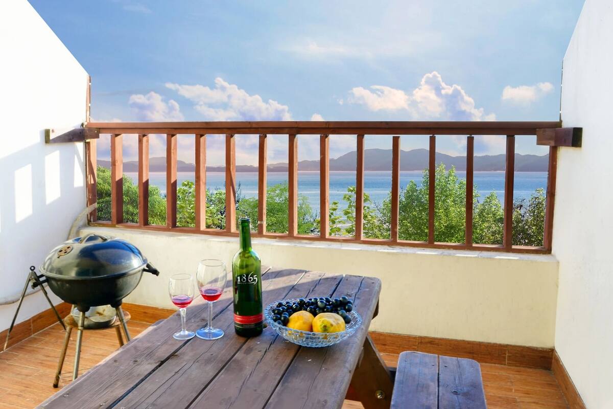 A balcony with a view of the sea, a grill, a bottle of wine, and a bowl of fruit.