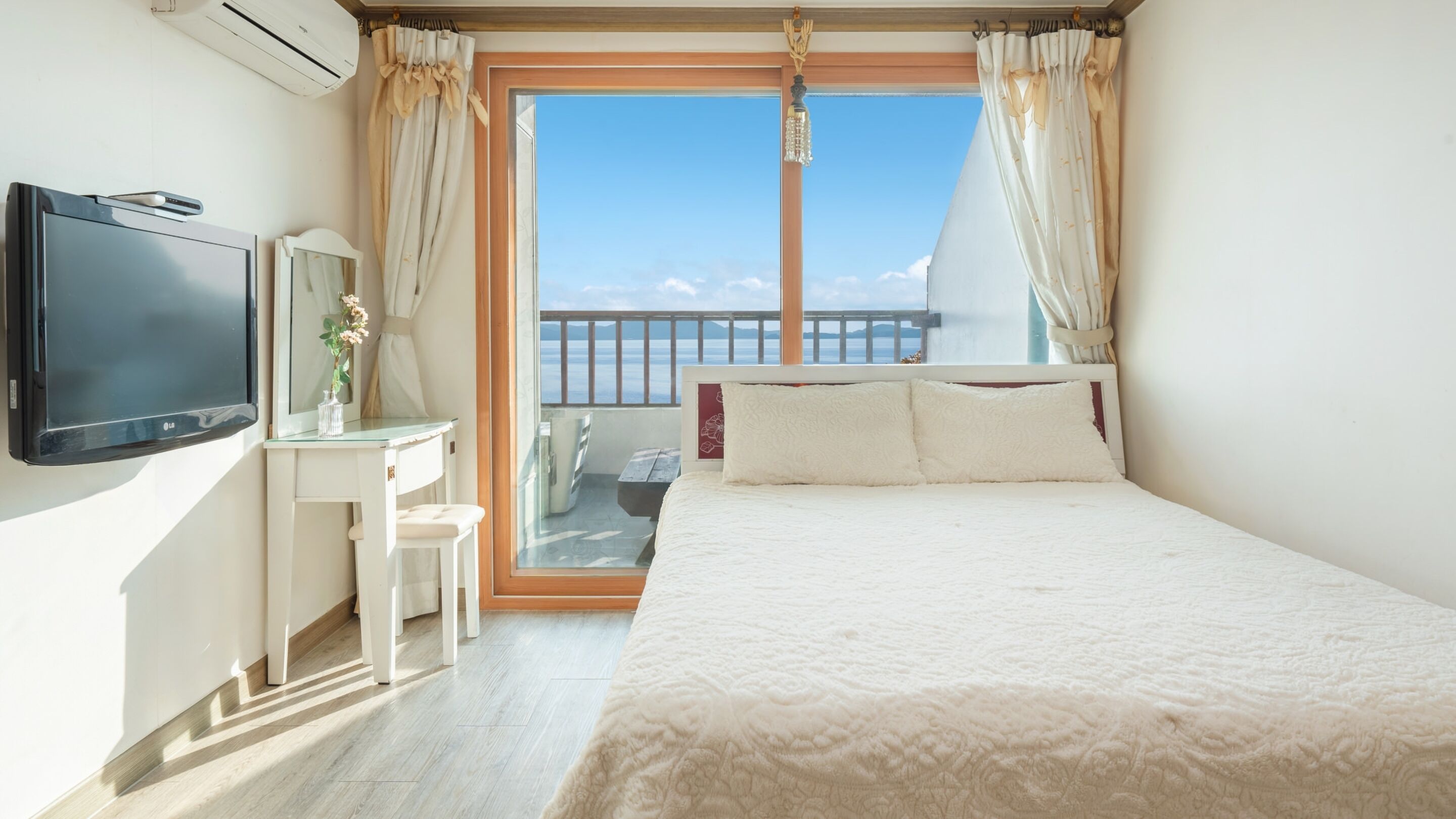 A bedroom with a bed, a desk, a TV, and a balcony with a view.