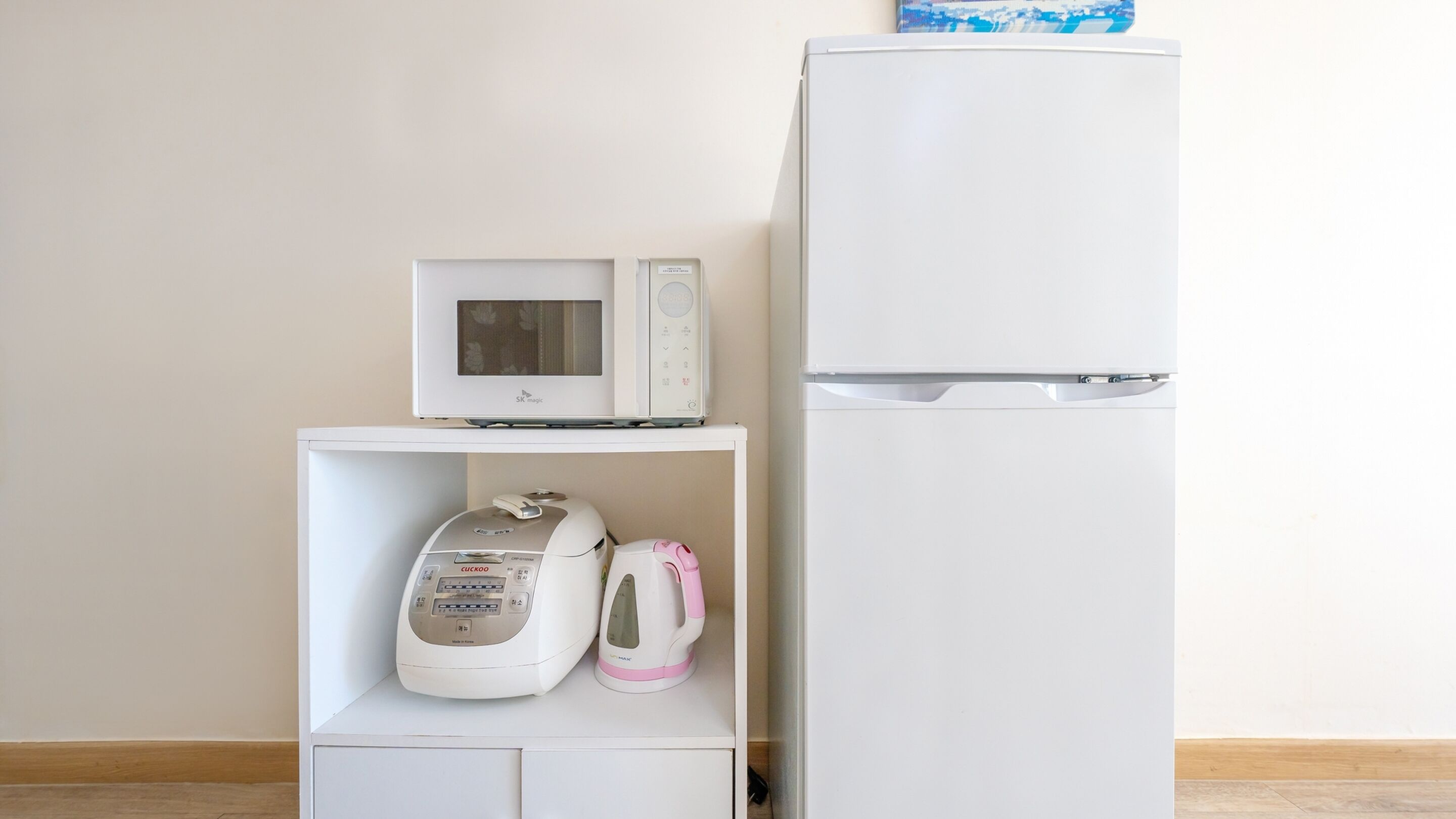 A white microwave on a shelf, a white kettle, and a white refrigerator in a kitchen.