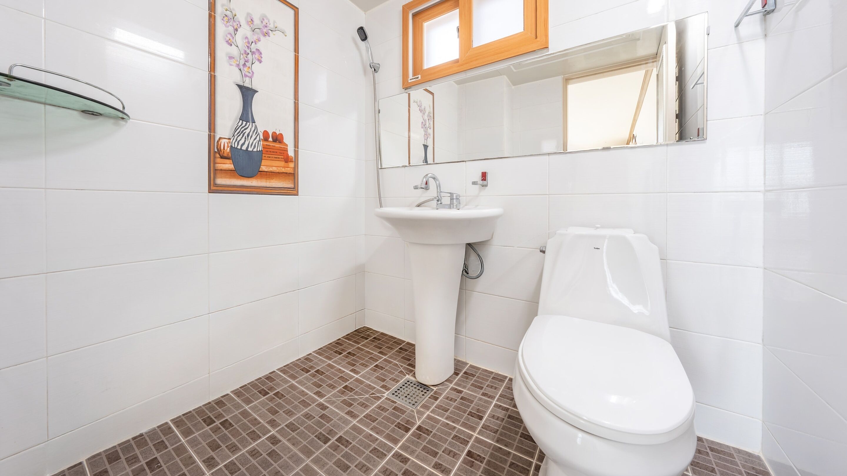 A bathroom with a white sink, a toilet, and a shower. There is a framed picture on the wall and a glass shelf.