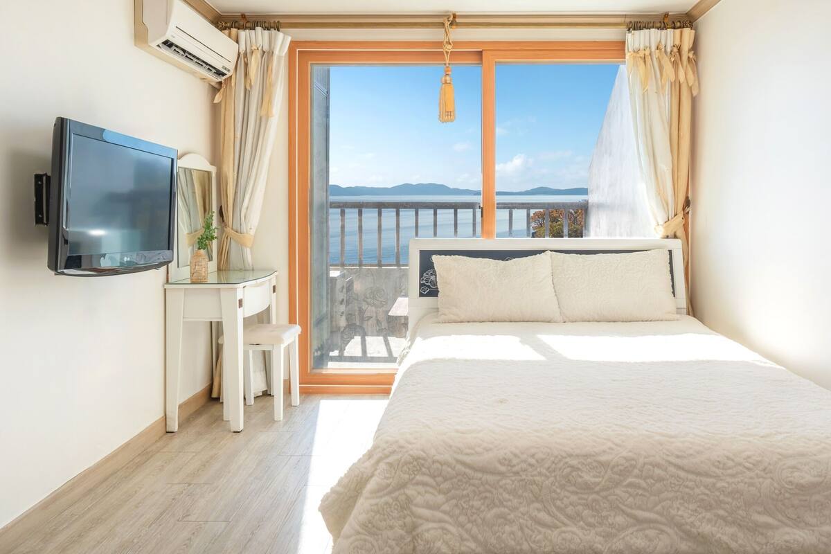 A bedroom with a bed, a TV, a desk, and a balcony with a view of the sea.