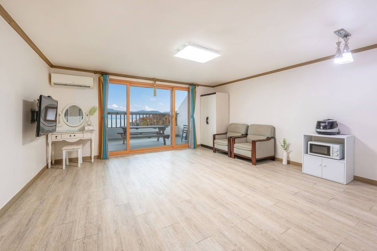A spacious room with a wooden floor, a sliding glass door leading to a balcony, a desk with a mirror, a sofa, and a kitchenette area.