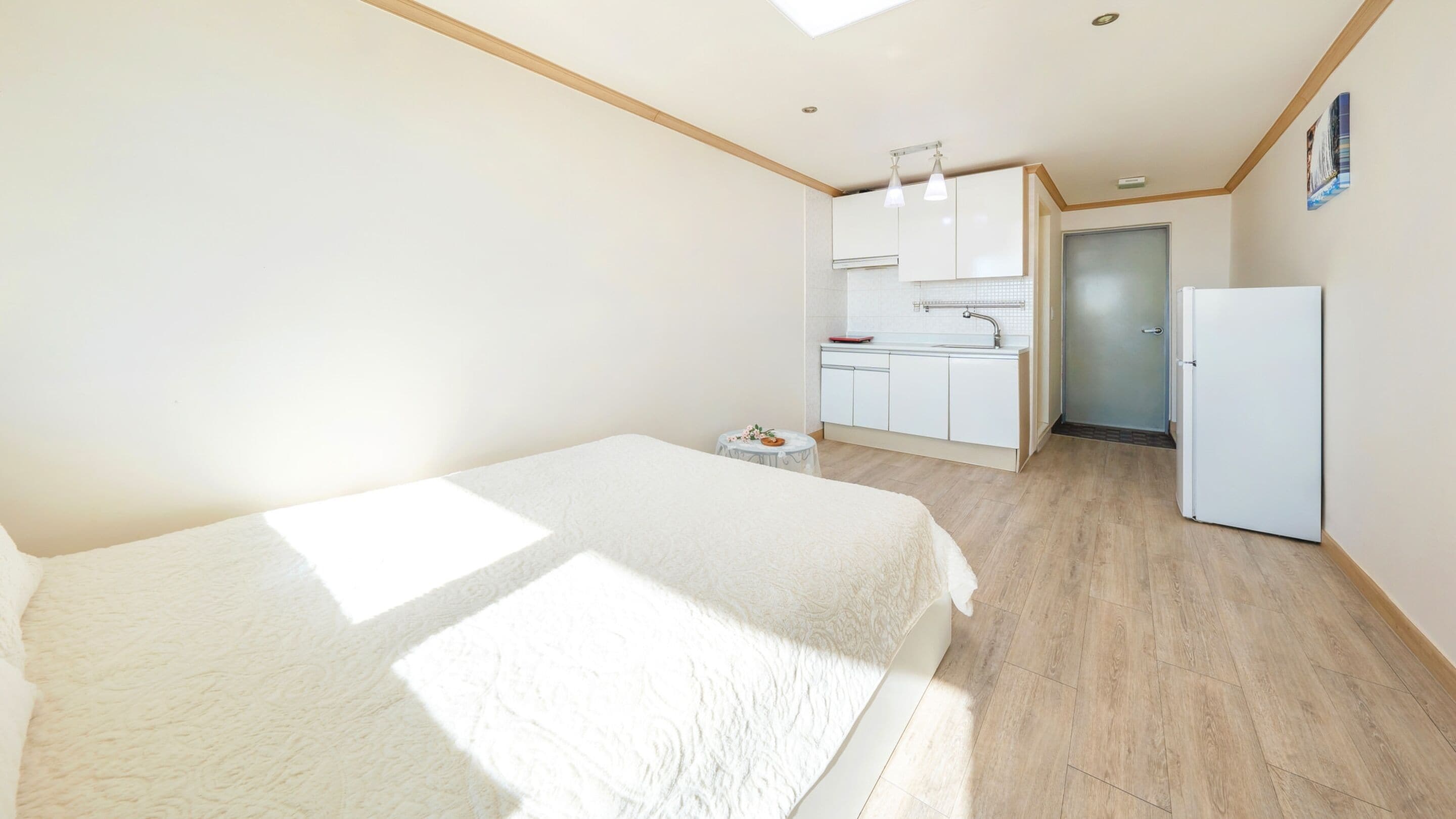 Un dormitorio con una cama, una mesita con un florero y una kitchenette equipada con refrigerador y armarios.