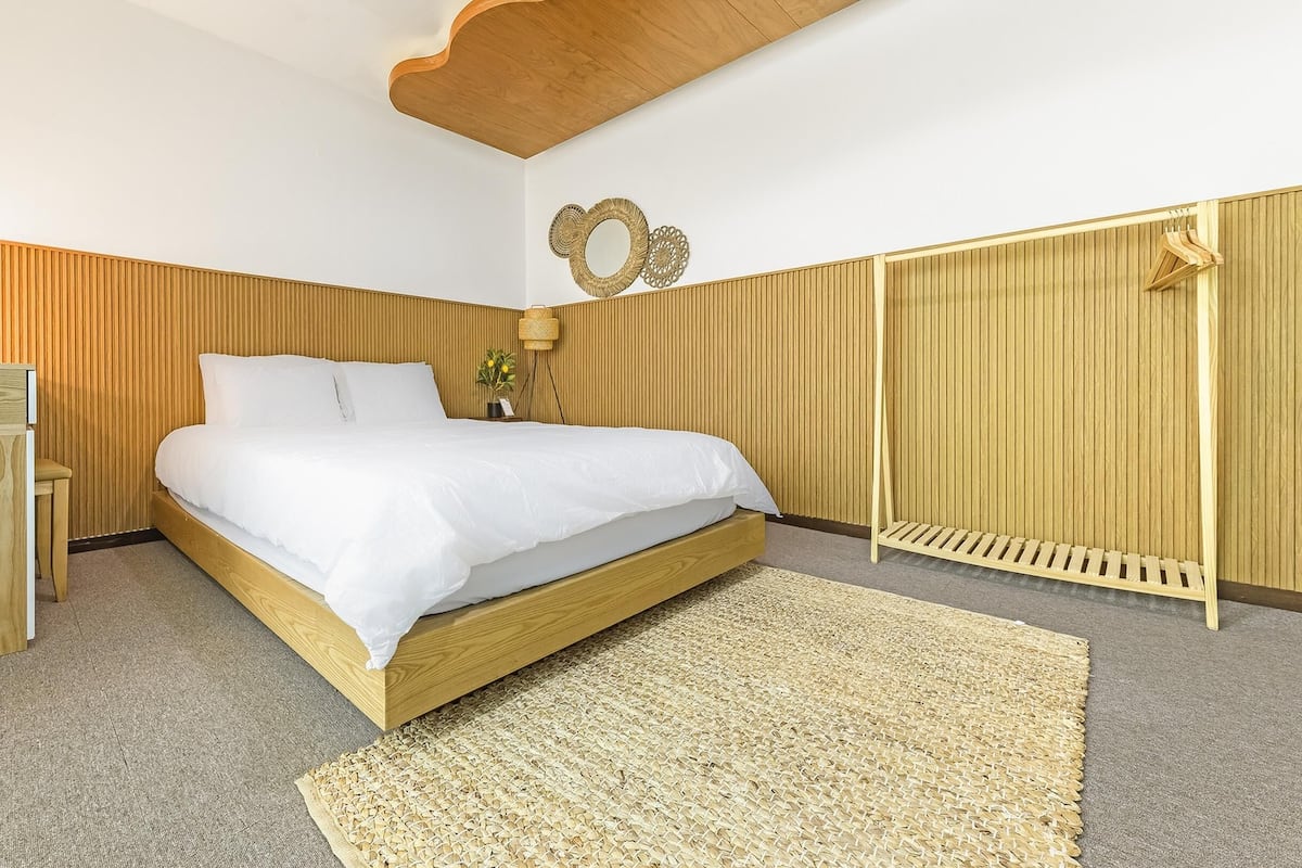 A modern bedroom with a wooden bed frame, a small bedside table, a wall-mounted mirror, and a wooden wardrobe.