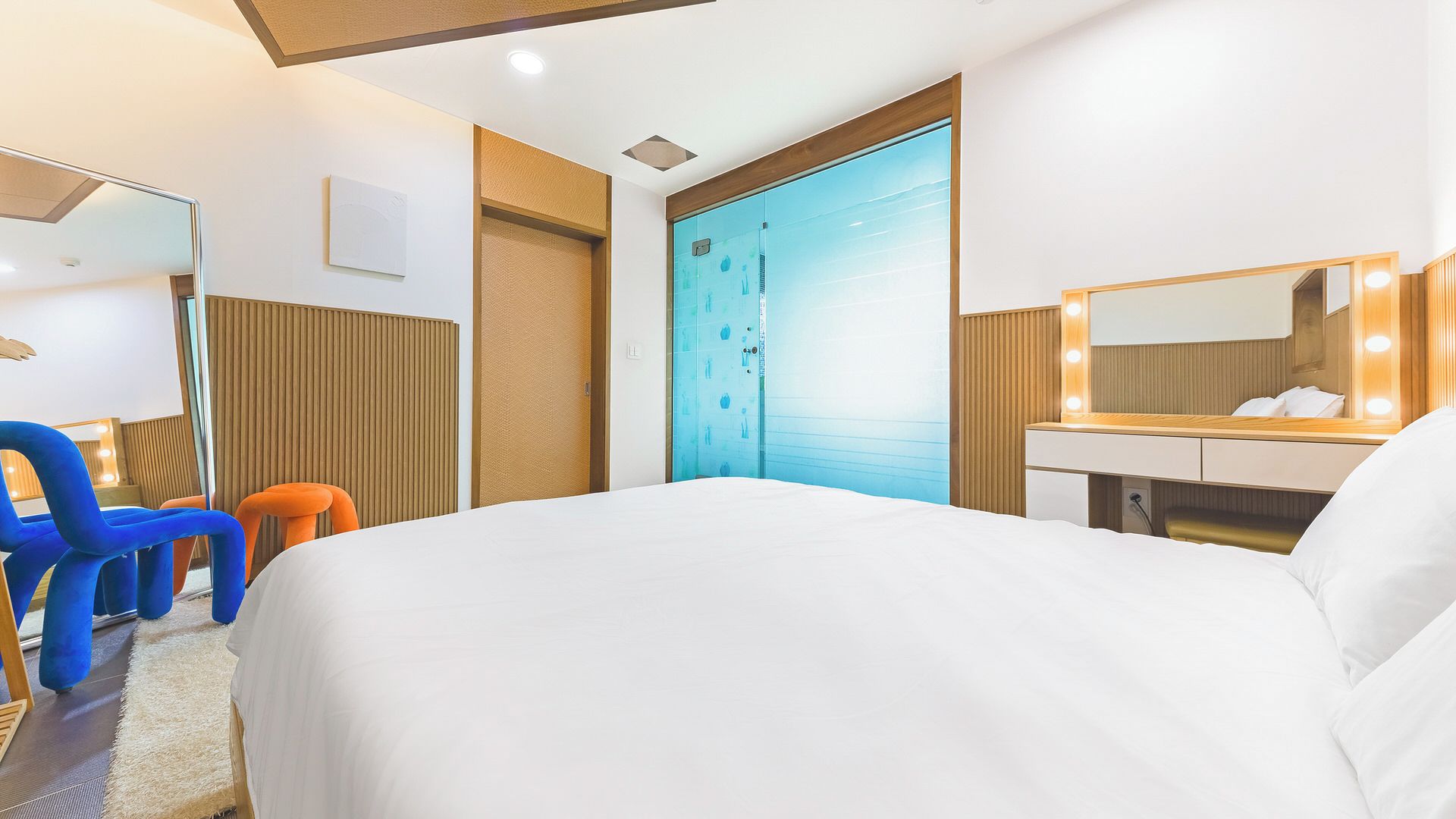 A hotel room with a bed, a vanity with a mirror and lights, a blue door, and a blue chair.