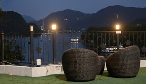 Un balcón con sillas de mimbre, baranda con diseños florales y vista a un lago con barcos y montañas a lo lejos.