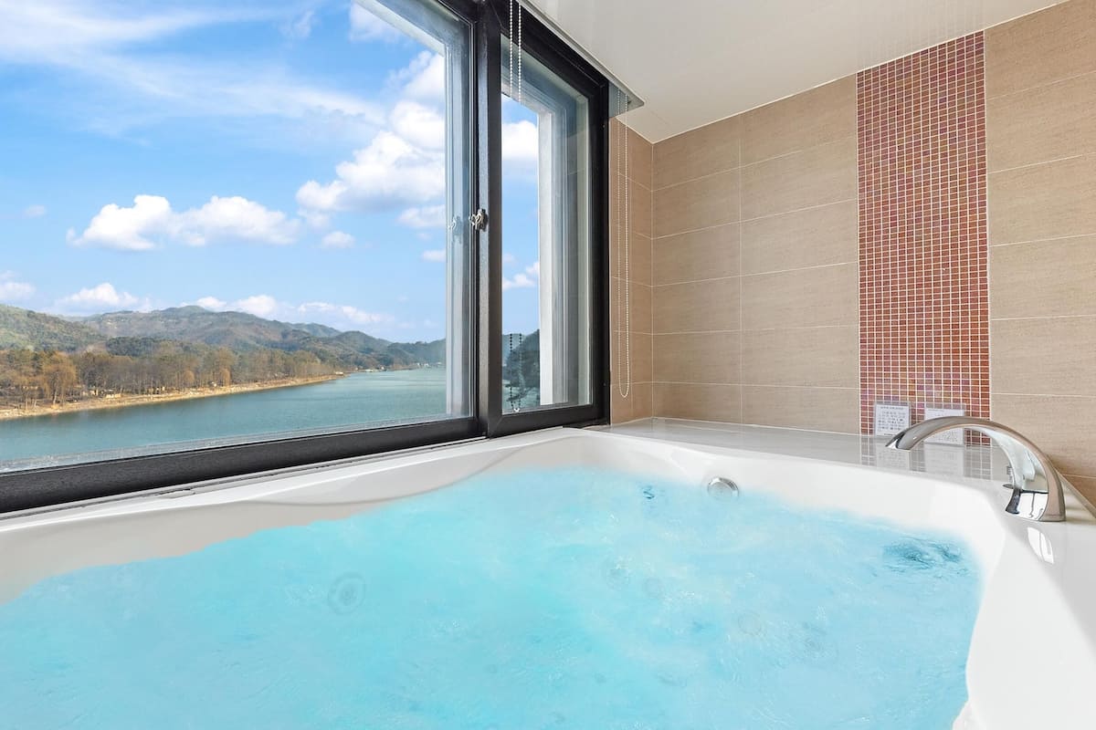 A bathroom with a large bathtub, a window with a view of a river and mountains, and tiled walls.