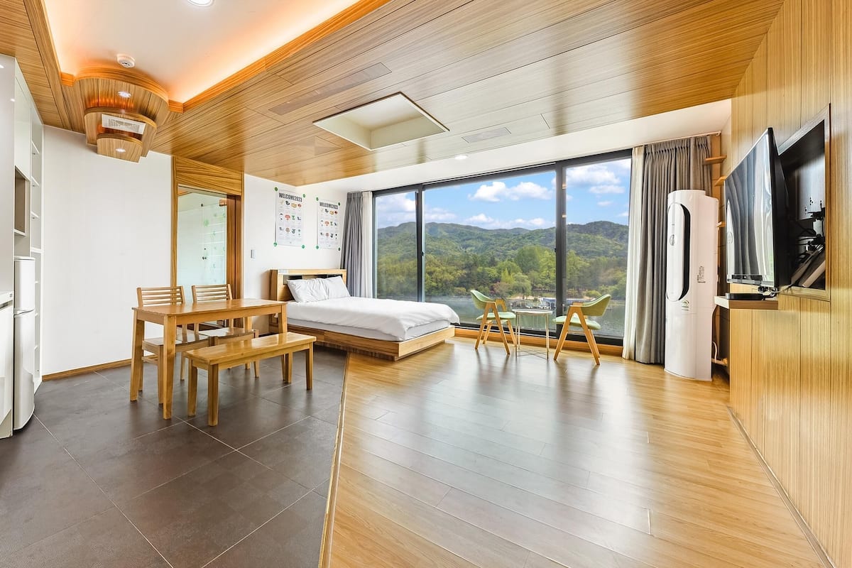 A modern hotel room with a wooden ceiling, a large window with a scenic view, a bed, a desk, and a TV.