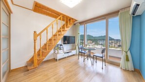 A spacious living area with a wooden staircase, a flat-screen TV, a dining table with chairs, and a large window offering a city view. - Samcheok Yedaum Pension (Samcheok)