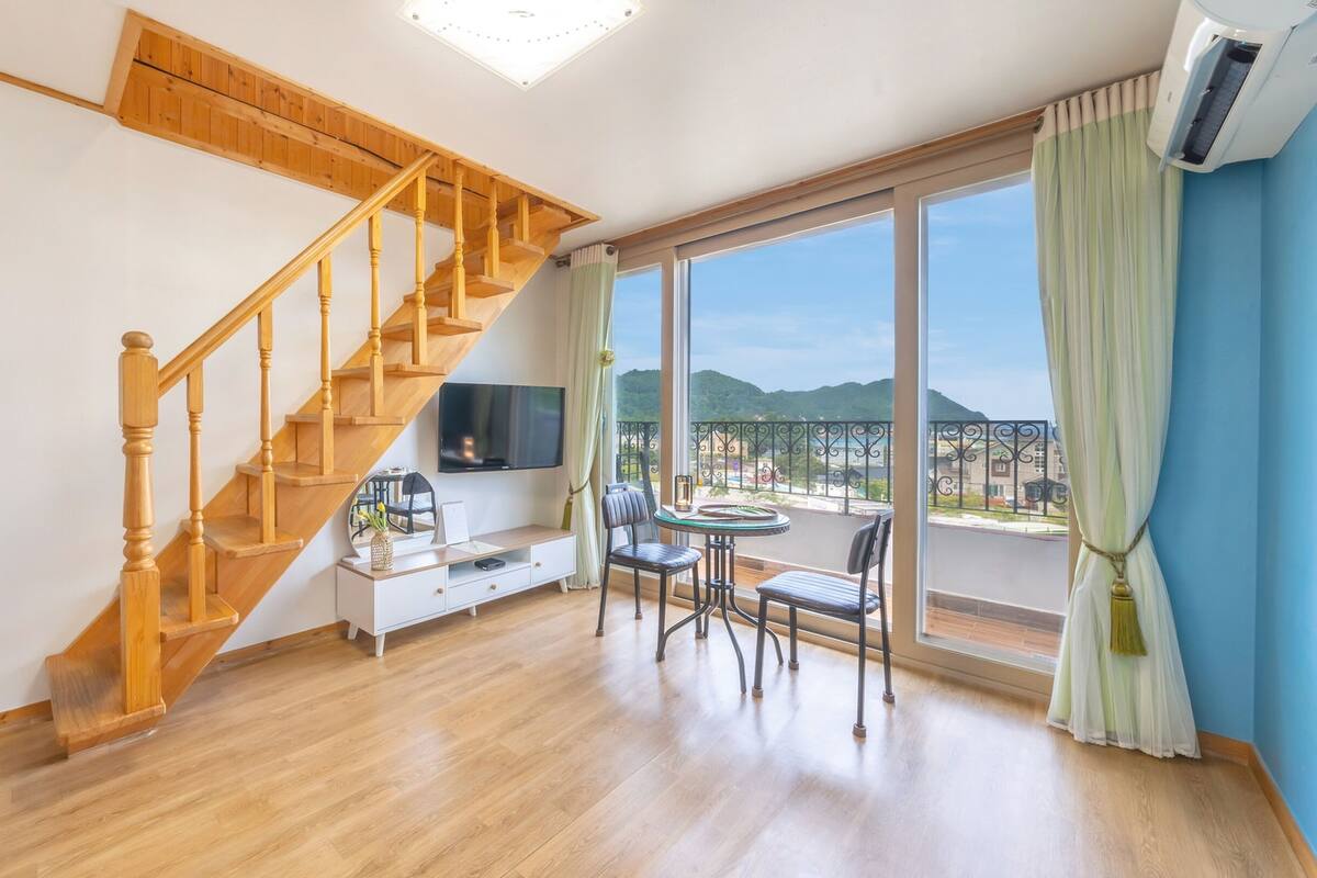 Un vaste espace de vie agrémenté d’un escalier en bois, d’un téléviseur à écran plat, d’une table à manger avec des chaises et d’une grande fenêtre offrant une vue sur la ville.