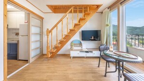 A spacious room with a wooden staircase, a dining area with a table and chairs, a flat-screen TV mounted on the wall, and a view of the outdoors through large windows. - Samcheok Yedaum Pension (Samcheok)