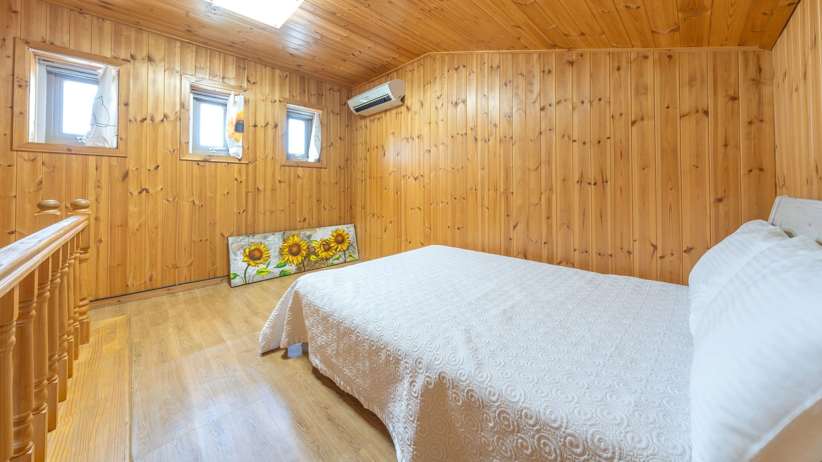 Habitación con paneles de madera, una cama, dos ventanas y un cuadro de girasoles.