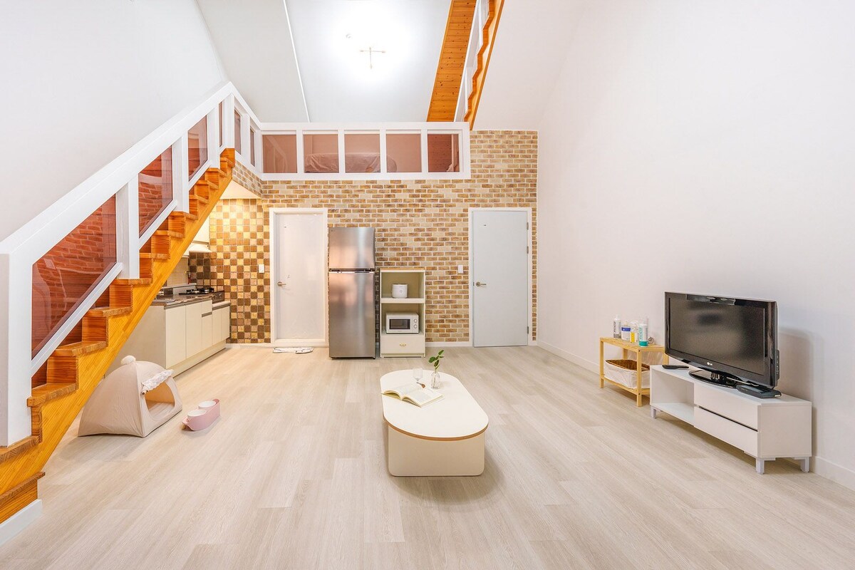 Un appartement moderne doté d’un escalier en bois, d’une cuisine avec réfrigérateur et four à micro-ondes, d’un coin repas avec une table et des chaises, et d’un téléviseur posé sur un meuble.