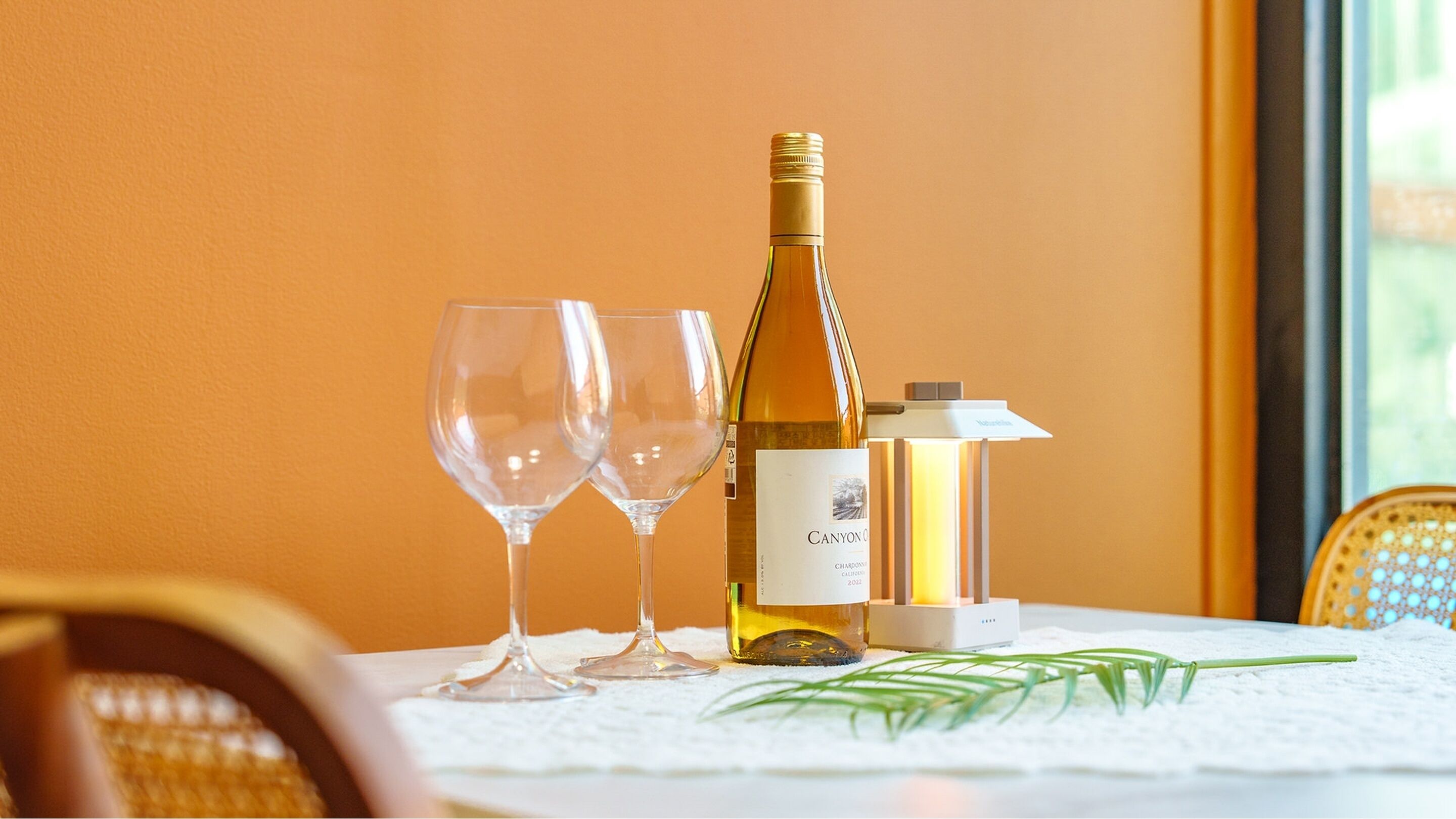 Een tafel met een fles Canyon Chardonnay en twee lege wijnglazen, een wijnwarmer en een kleine plant.