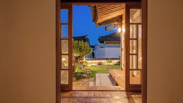 Una puerta de madera abierta que da a un jardín japonés tradicional, con un suelo de madera y vistas a un edificio con tejado de tejas.