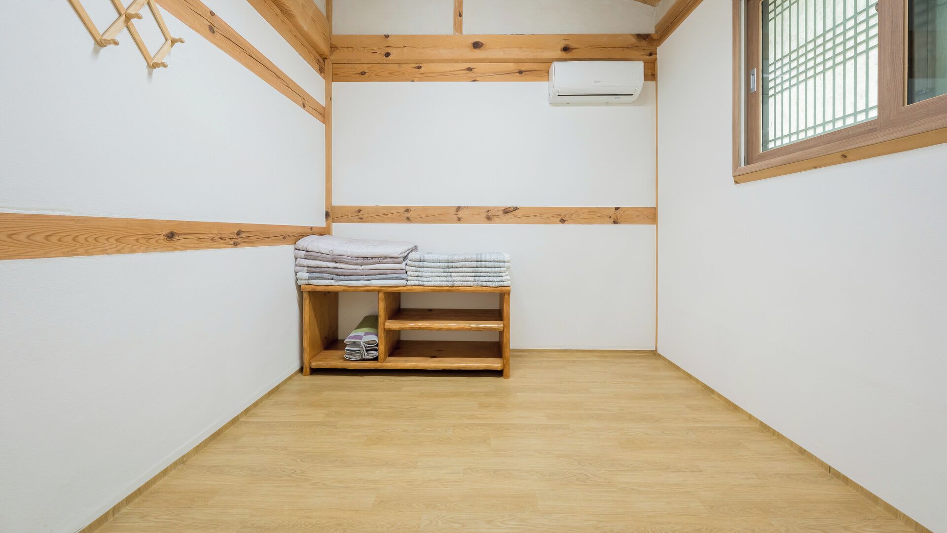 A small, clean room with a wooden bench, a wall-mounted air conditioner, and a window with a grid pattern.