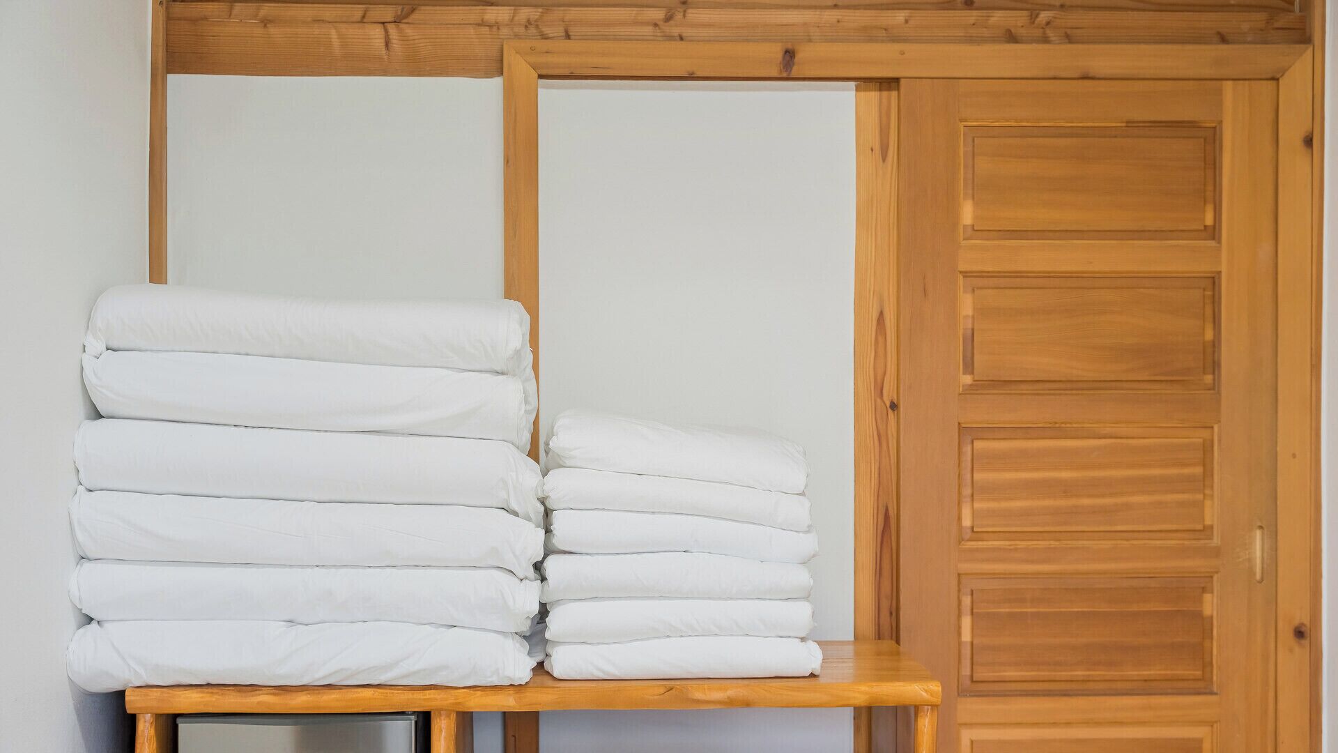 Una pila de ropa de cama blanca sobre una mesa de madera, frente a una pared de madera con una puerta.