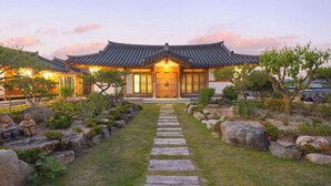 Un edificio de estilo coreano tradicional con una puerta de madera, un sendero de piedra que conduce a la entrada y un jardín con rocas y arbustos.